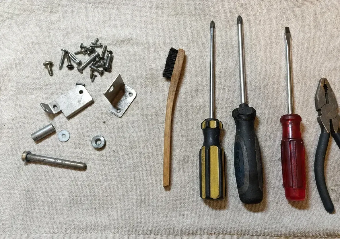 Overhead view of loose screws and small steel brackets beside a wire brush, three screwdrivers and a pair of pliers on a cream drop cloth