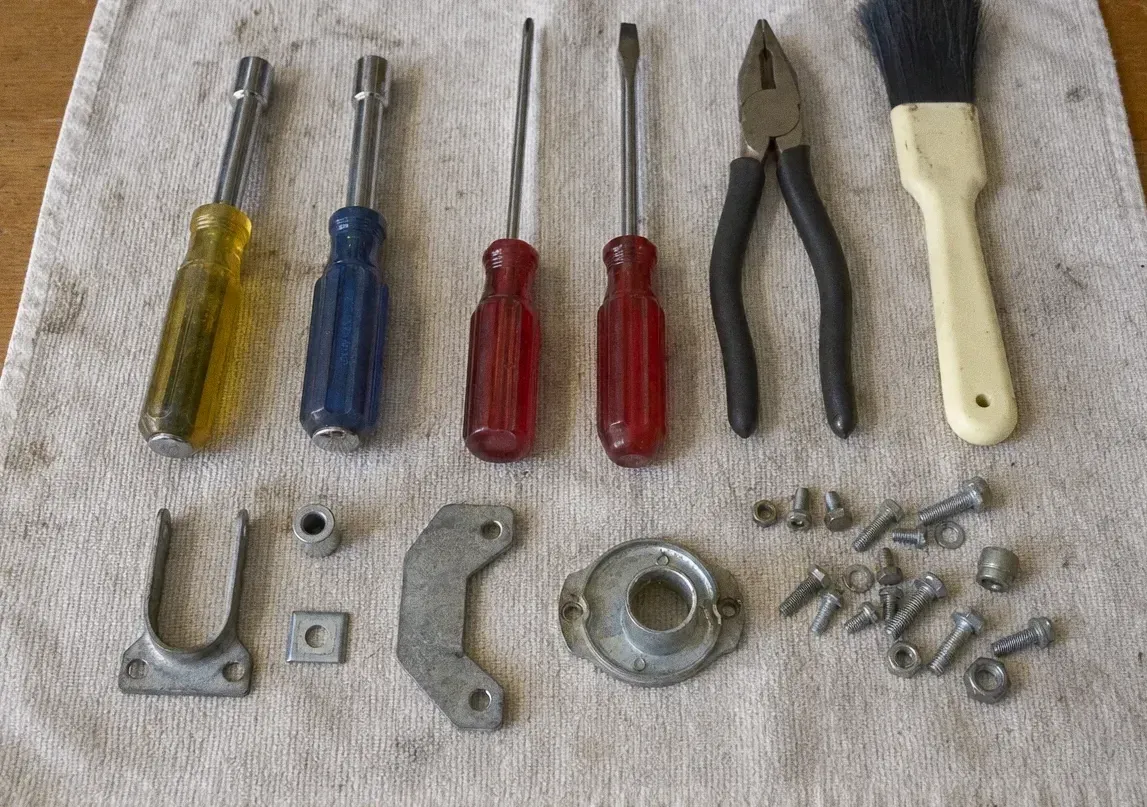 Overhead view of four screwdrivers and pliers laid beside a U-bracket, a cam plate, a bearing collar and a row of bolts and nuts