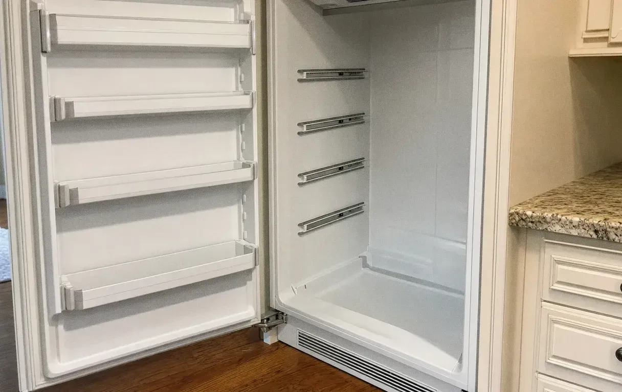 A white built-in refrigerator with both doors standing open on empty shelf rails, wood floor and cabinetry either side